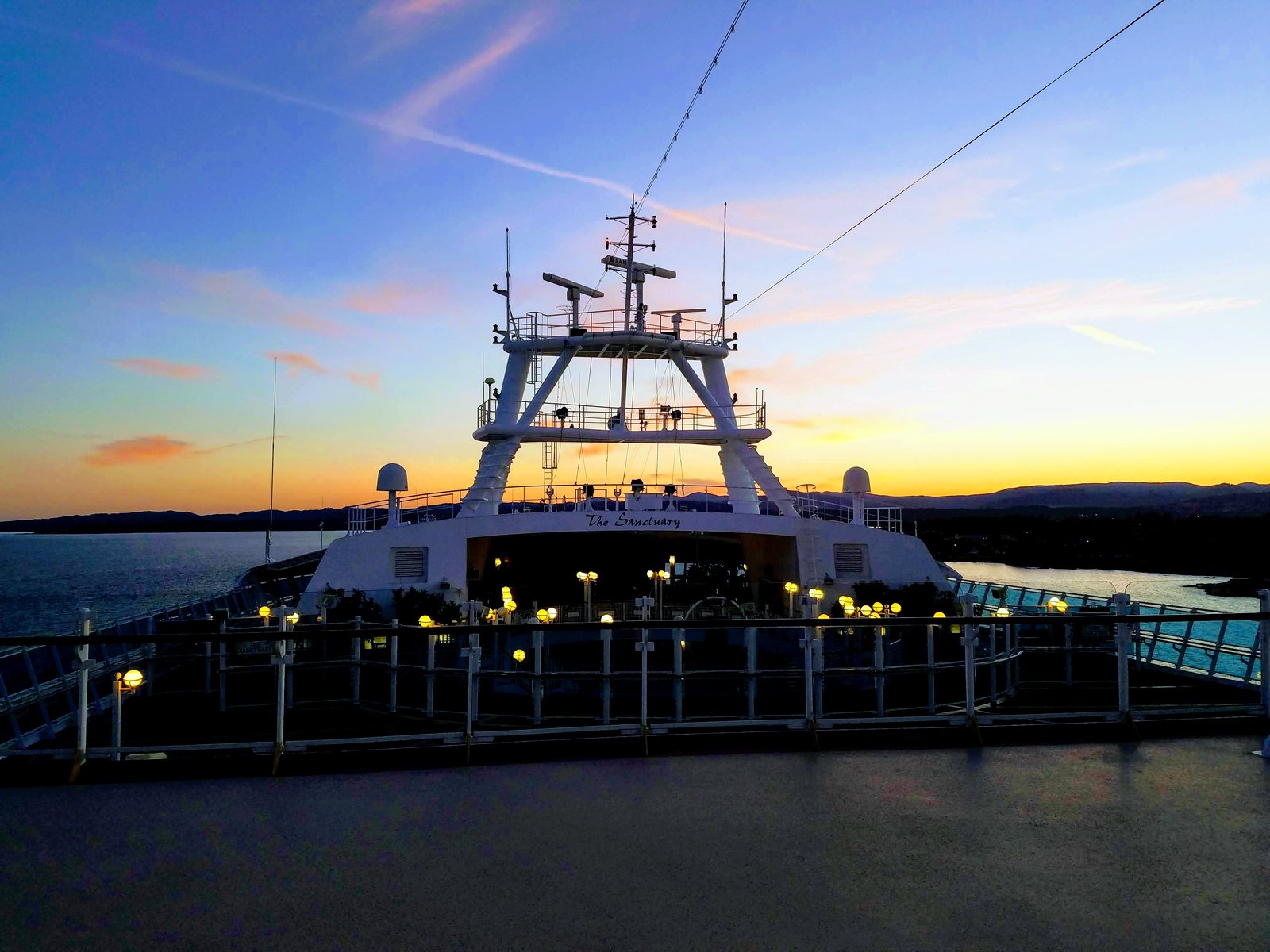 The forward decks and mast of a cruise ship lit up at dusk under a pink and blue sky, approaching Victoria