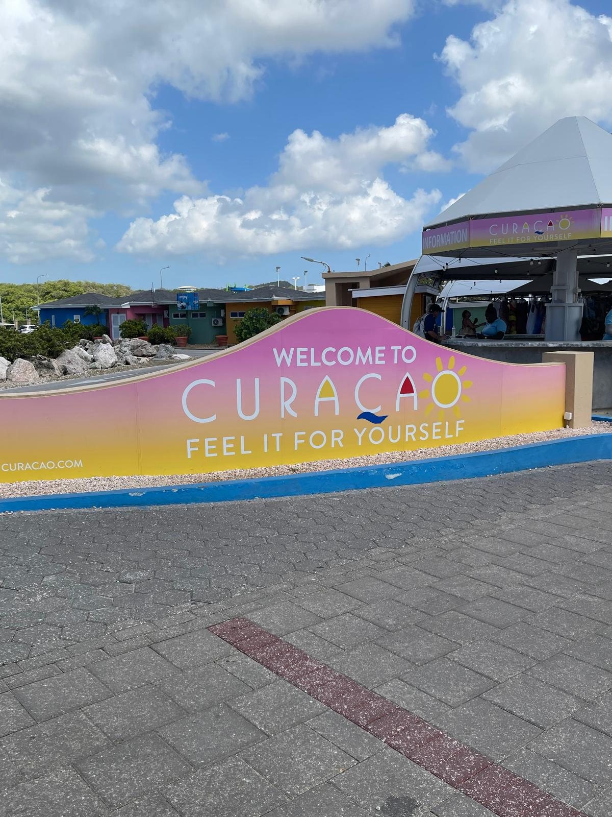 A bright painted WELCOME TO CURACAO sign on the pier walkway, with the words Feel It For Yourself beneath