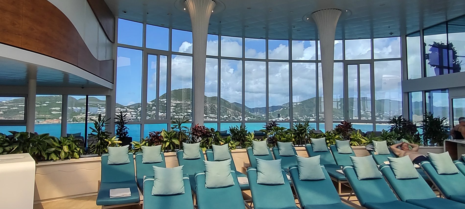 A quiet ship solarium, rows of turquoise loungers under a curved wall of windows looking out at an island