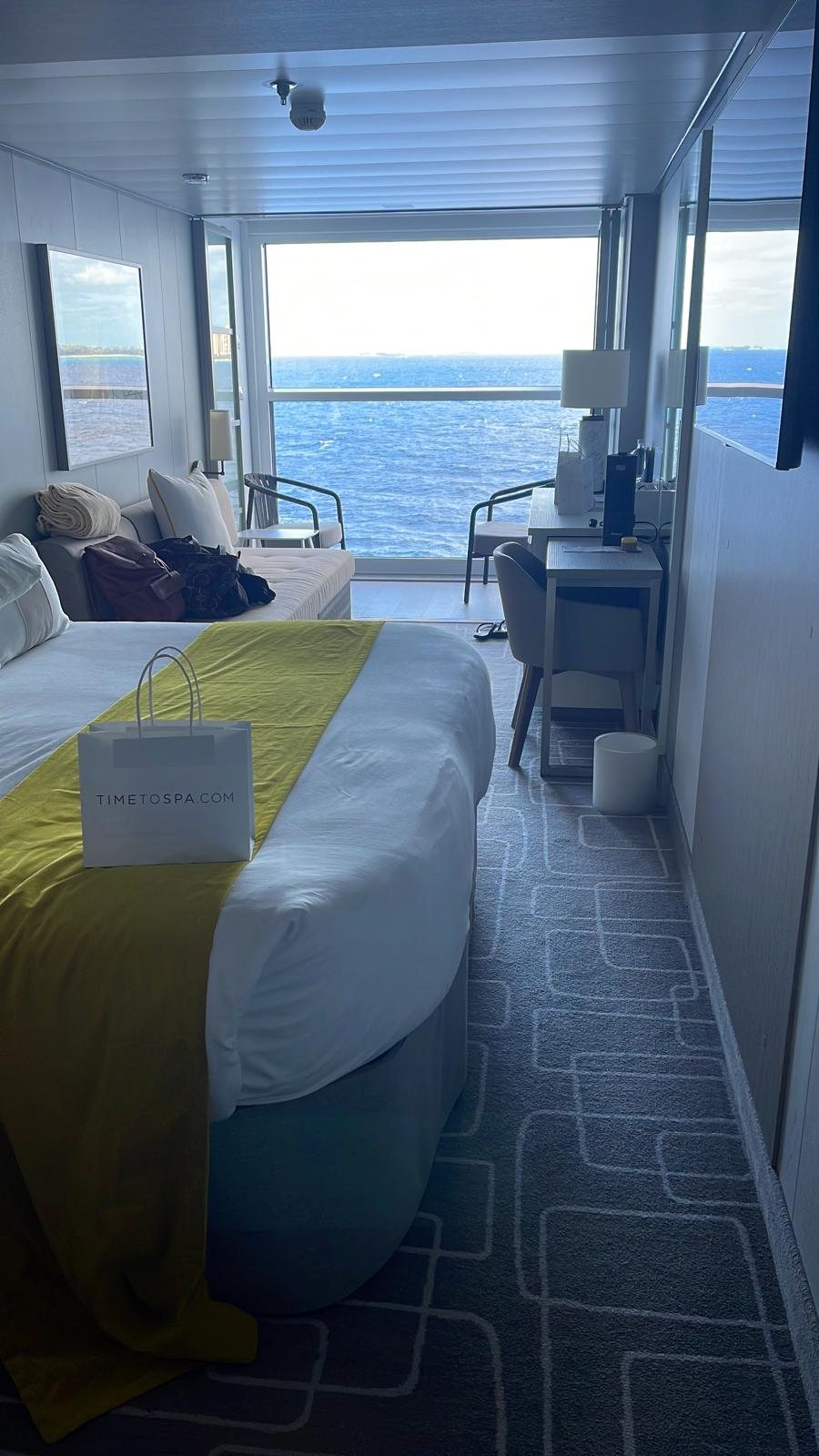 A cruise ship stateroom with the balcony door open, a made bed in the foreground and blue sea beyond